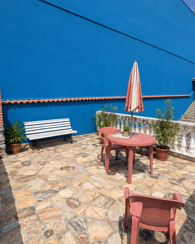 Terraço com mesa, cadeiras e guarda-sol fechado entre plantas, de frente para o muro azul