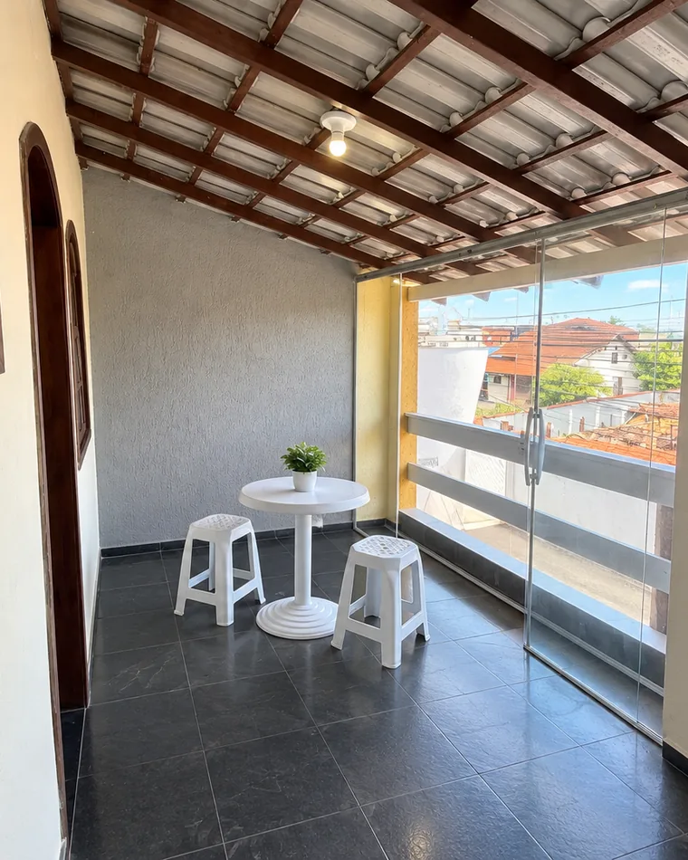 Varanda com mesa e banquetas brancas sob telhado de madeira, com vista para os telhados do bairro