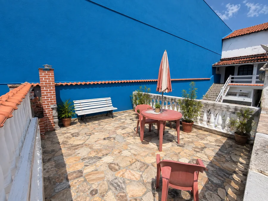 Terraço ao ar livre com mesa, cadeiras e guarda-sol entre plantas, com muro azul e céu limpo ao fundo