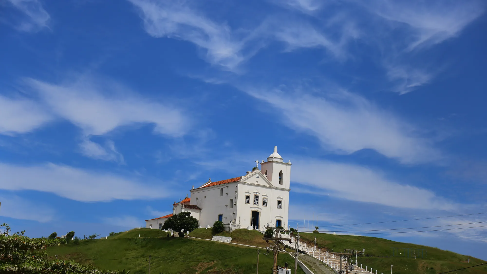 A Igreja de Nossa Senhora de Nazareth, branca, no alto de uma colina verde sob um grande céu azul com nuvens finas