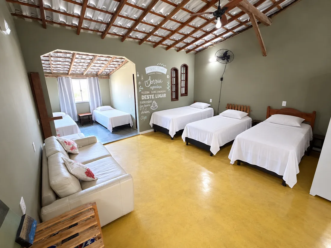 Quarto compartilhado amplo dividido em ambientes, com camas de solteiro, sofá e telhado de madeira aparente