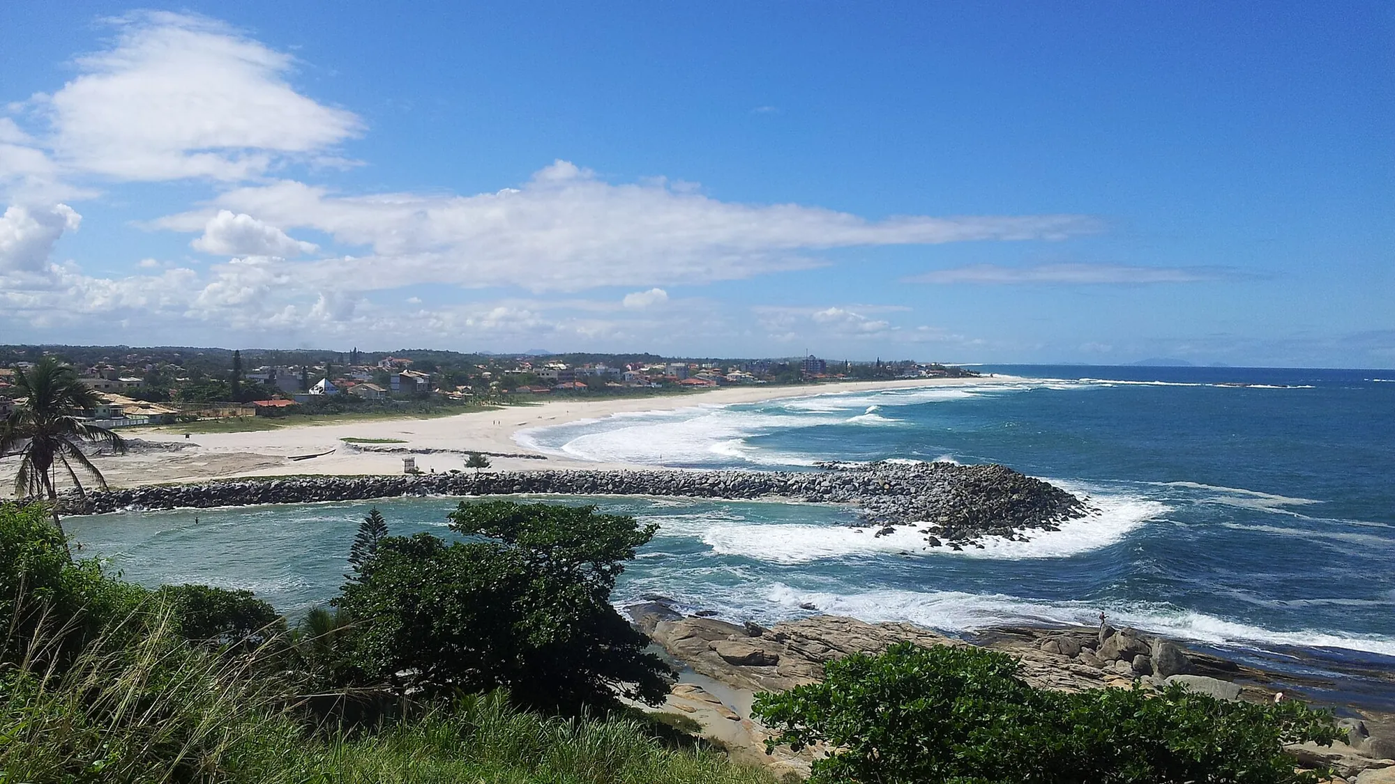Panorama de Saquarema: a longa faixa de areia curvando até os morros, o molhe de pedras avançando no mar e as ondas quebrando sob céu azul
