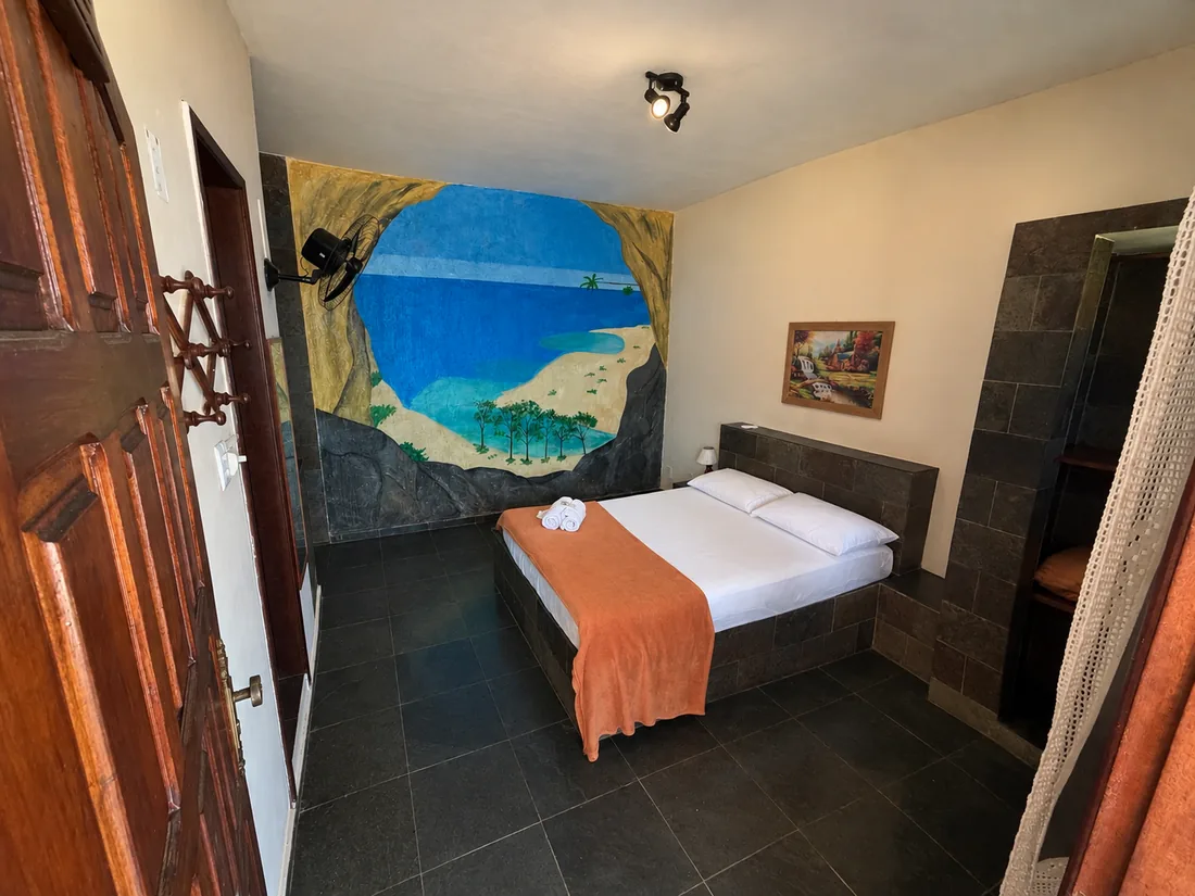 Suíte com cama de casal e manta laranja, parede com mural pintado de praia e piso de ardósia