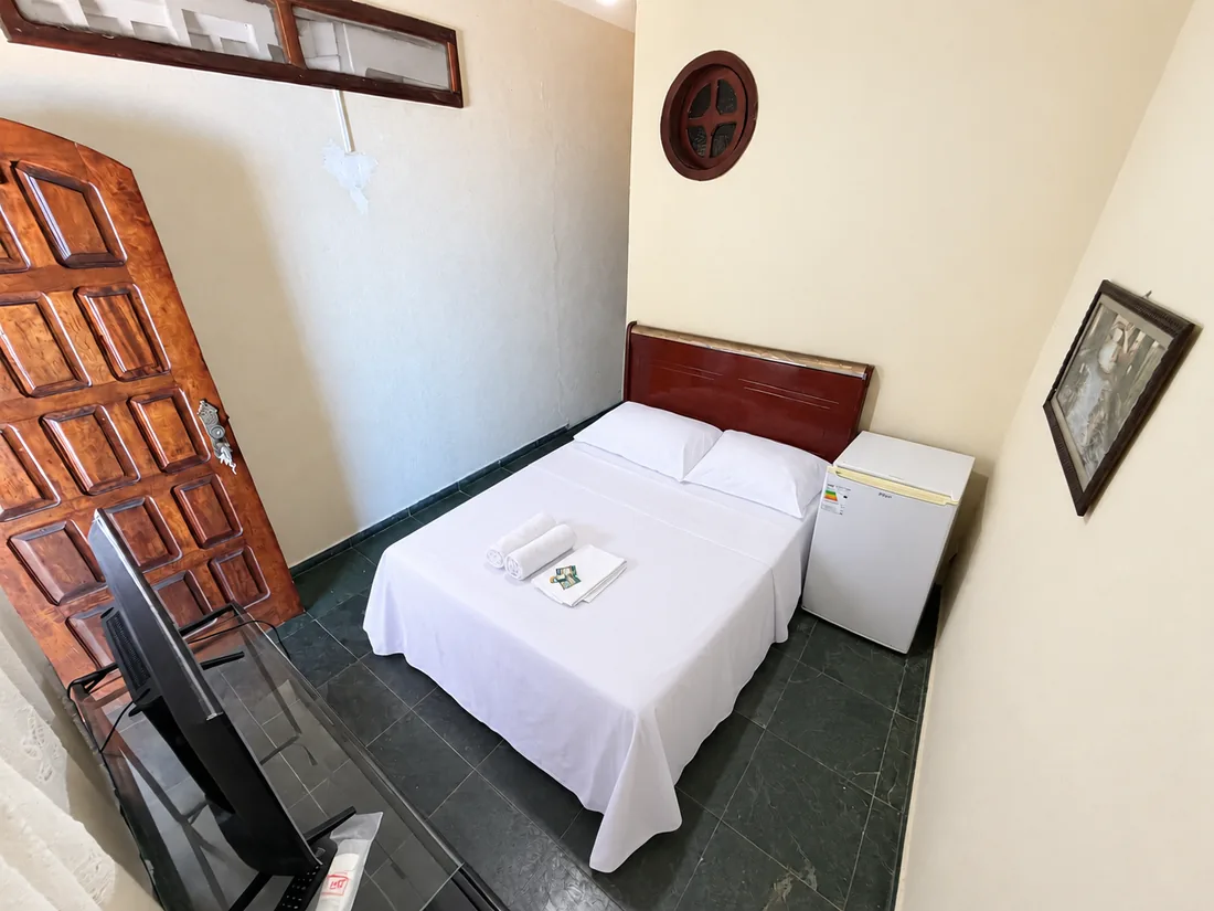 Suíte com cama de casal, frigobar branco ao lado e porta de madeira maciça